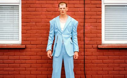 Daytime image of a man in a light blue suit with tie fastening against a red brick wall, white framed windows with white venetian blinds either side