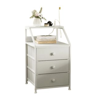 A white nightstand with three drawers and books and flowers on top of it