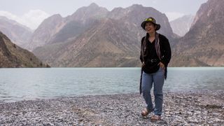 A woman wearing Patagonia Women’s Chambeau Rock Pants stands at the edge of a lake, with mountains in the background.