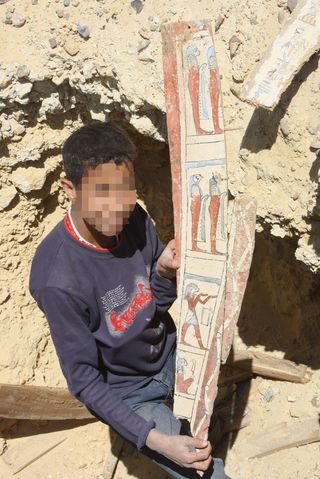 A child working in a pit holds up a prized find, a wooden board, likely from an ancient coffin, which contains a well preserved painting.