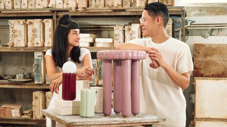A couple of designers dressed in white workwear with short-sleeved tops and trousers leans onto a set of colorful homeware creations, including a pink stool, a green and a bordeaux light in a wood-crammed studio illuminated by natural light.