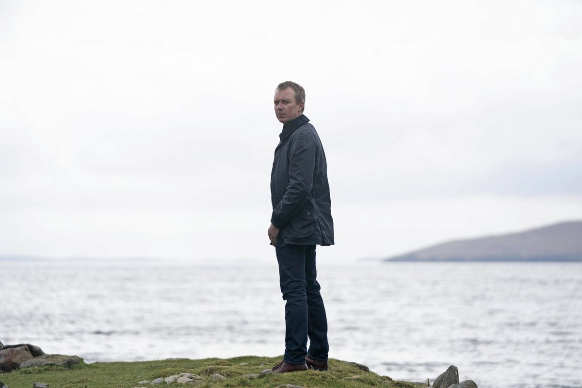 Shetland&#039;s DC Sandy Wilson (Steven Robertson) stands on a cliff edge with the ocean behind him. His body is turned away from the camera but he is looking back over his shoulder at us.