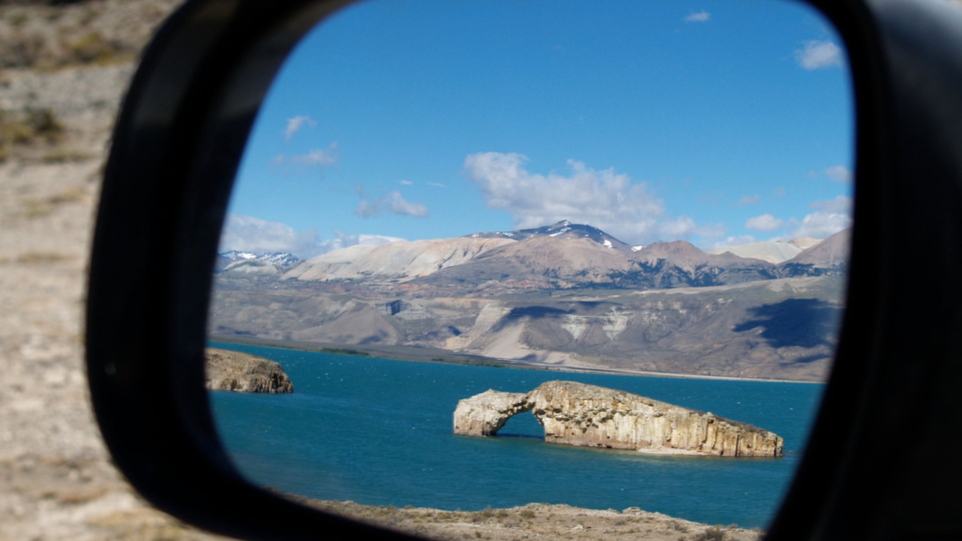A photo of a car wingmirror showing Lake Posadas and its famous rocky arch reflected in the mirror