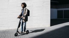 A man riding one of the best electric scooters down the street, there's a white brick wall behind him