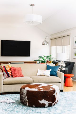 beige living room with beige and black couches, tv, bright cushions, cowhide footstool, blue textured rug, silver lamp, orange stool, blinds