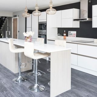 White kitchen with white cabinets, island with bar stools and grey wooden flooring