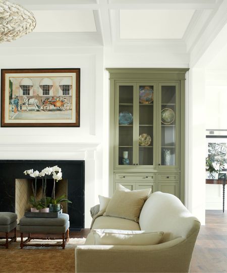 Transitional living room with white walls, large fireplace, a built-in cabinet painted in Rosemary Sprig by Benjamin Moore, a cream sofa, Persian rug, and a stool holding an orchid.