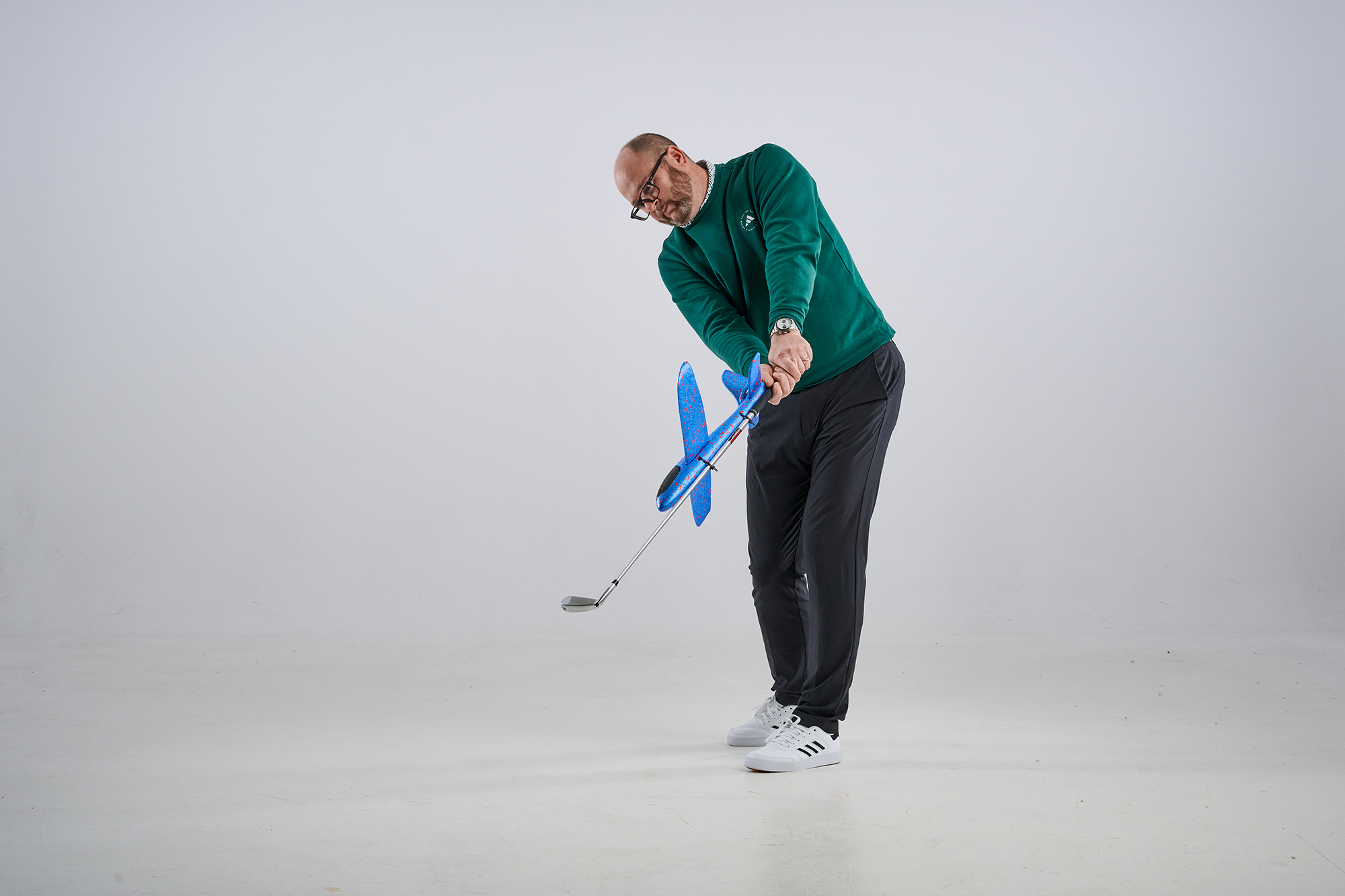 Russell Covey just past impact position in an iron shot, with a toy plane attached to the shaft of the iron as part of a ball striking drill