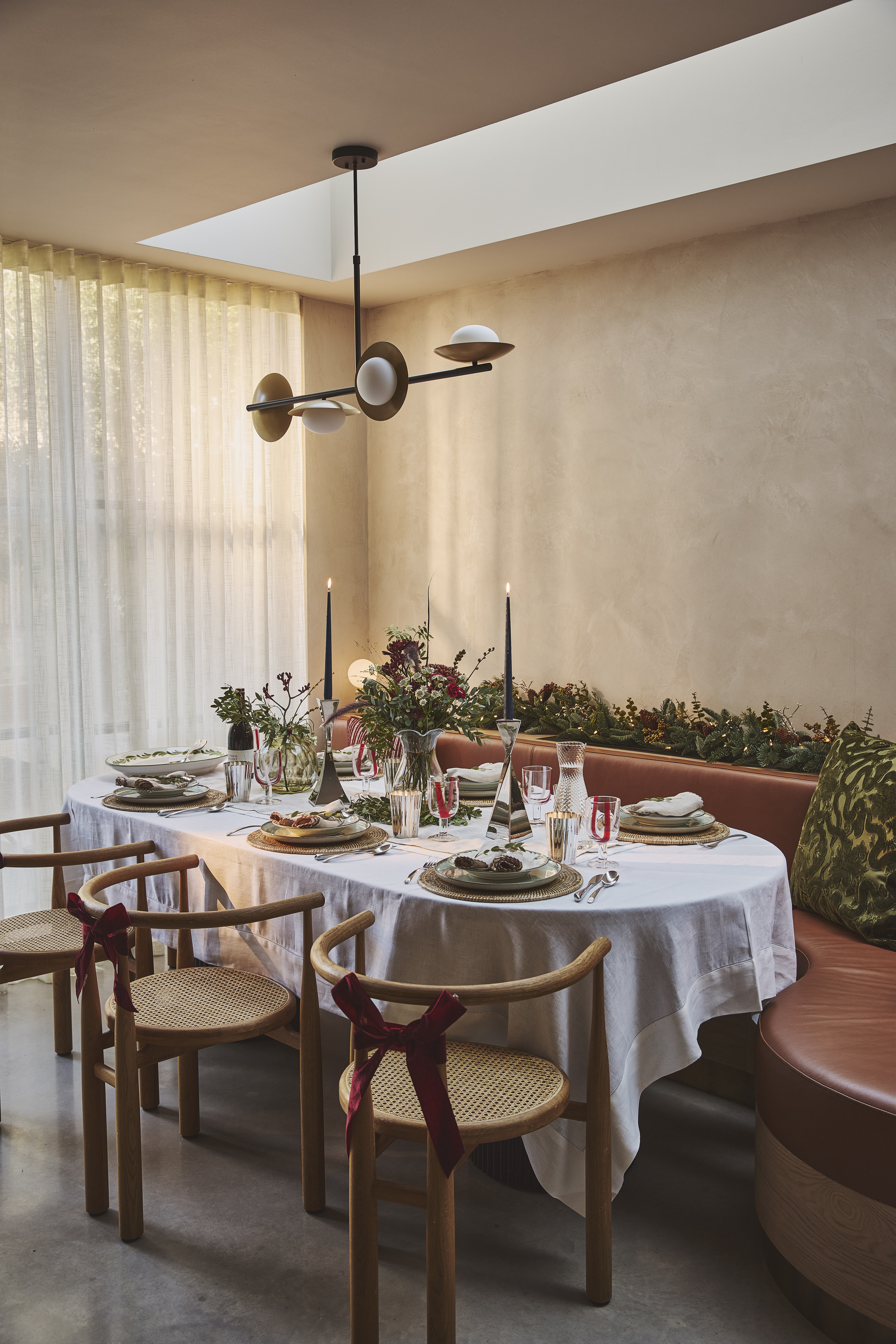 a christmas table with banquette seating decorated with lit festive foliage on one side and wooden dining chairs with red velvet bows on the back on the other with neutral limewash walls, and sheer curtain-covered windows in the background with a pendant ceiling light hanging above the set table with lit candles
