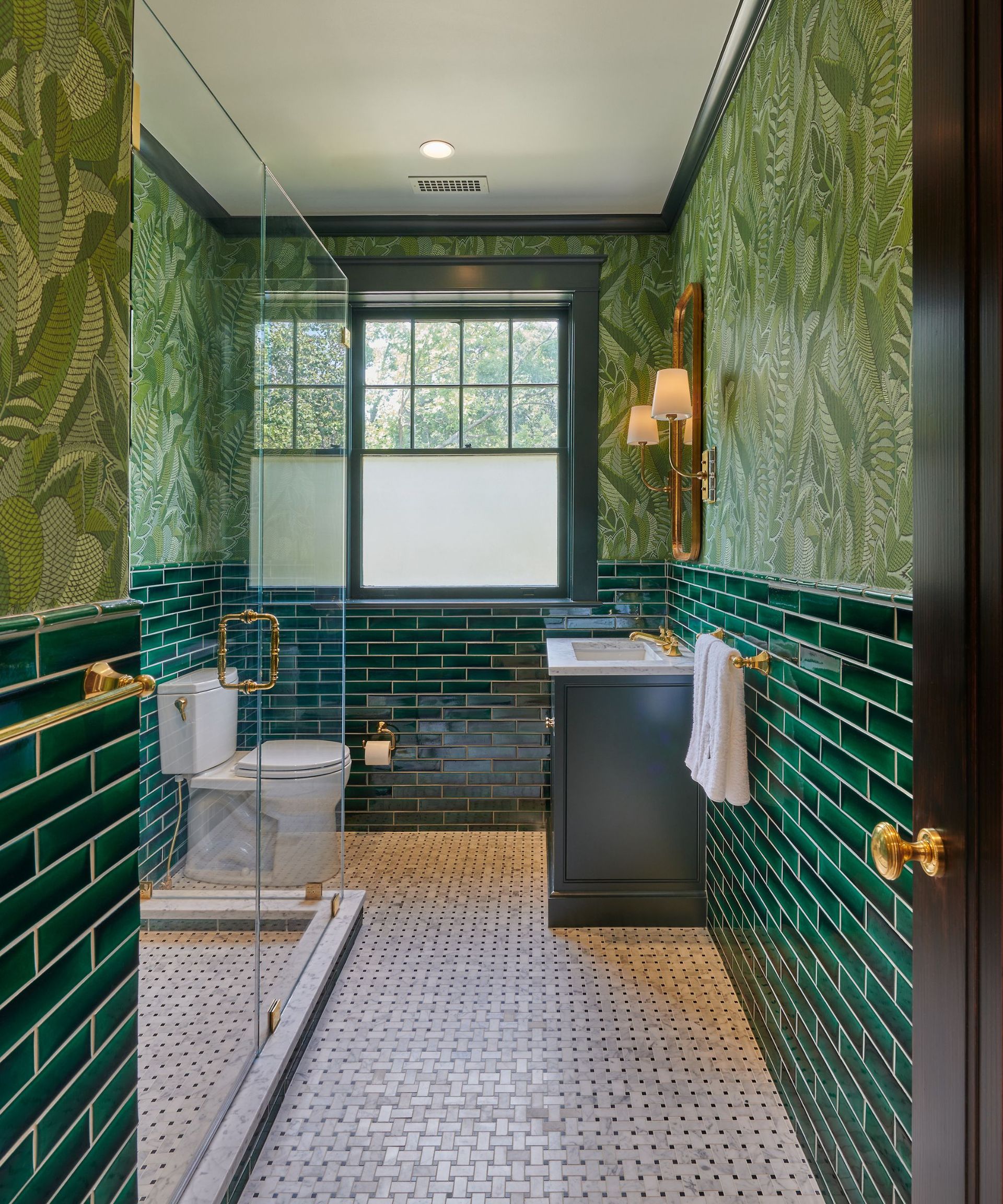 A small green bathroom with dark green subway towels on the lower half of the walls and a leaf-themed wallpaper on the upper walls.