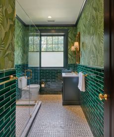 A small green bathroom with dark green subway towels on the lower half of the walls and a leaf-themed wallpaer on the upper walls. 