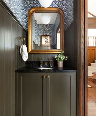 A small bathroom with dark brown wooden shiplap and vanity, a curved gold mirror, and blue and white patterned wallpaper