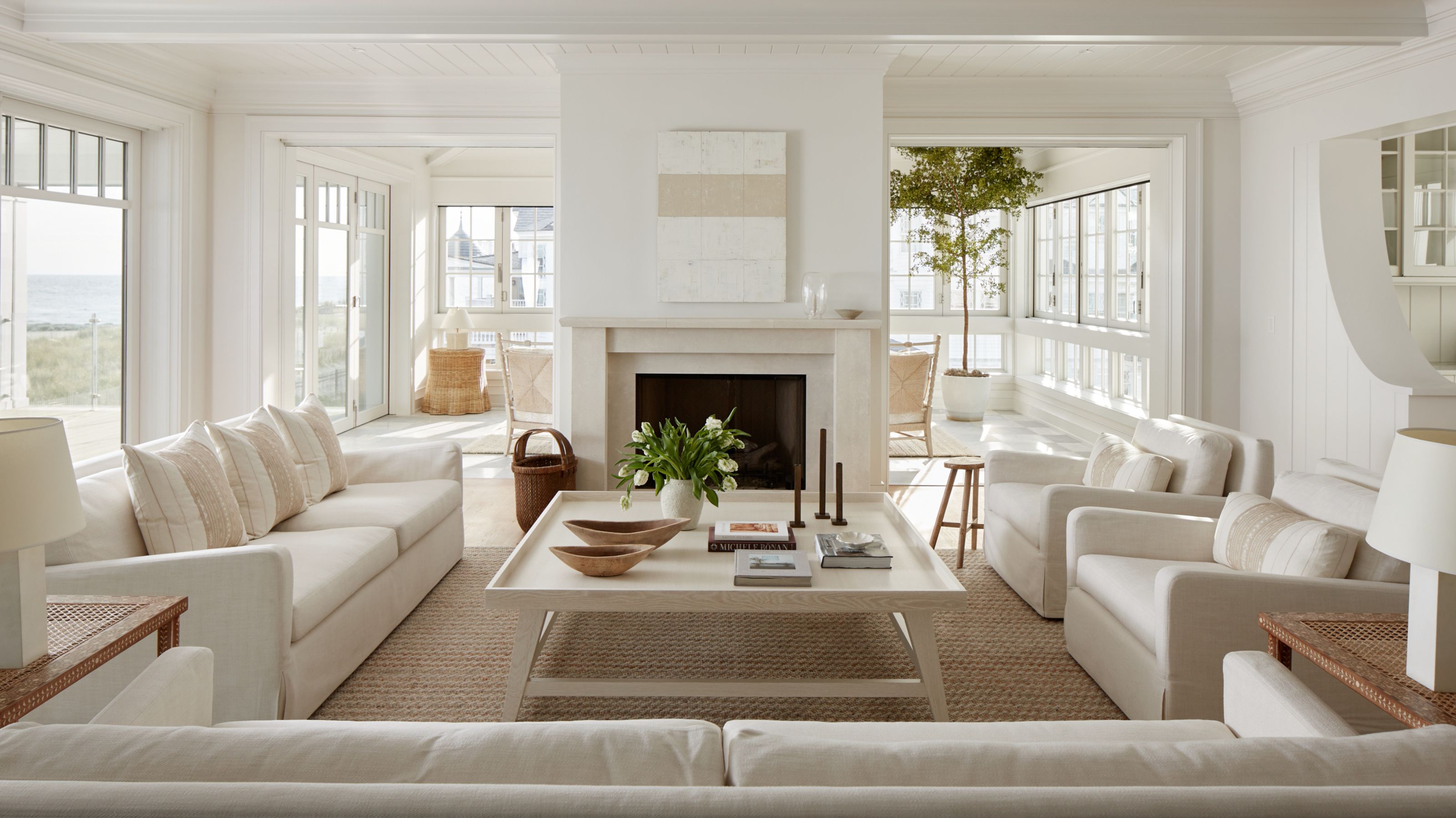 An expansive, light-filled living room featuring symmetrical cream sofas, a large square coffee table, and a central stone fireplace. The space is anchored by a natural jute rug and offers views of a sunroom and the coast through large windows.