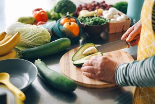 Woman cutting the stone out of an avocado, surrounded by other fruits and vegetables to meal prep with