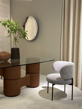 A gray chenille chair with black hardware beside a glass table with a brown stand in a beige room with a silver reflective plate on the wall