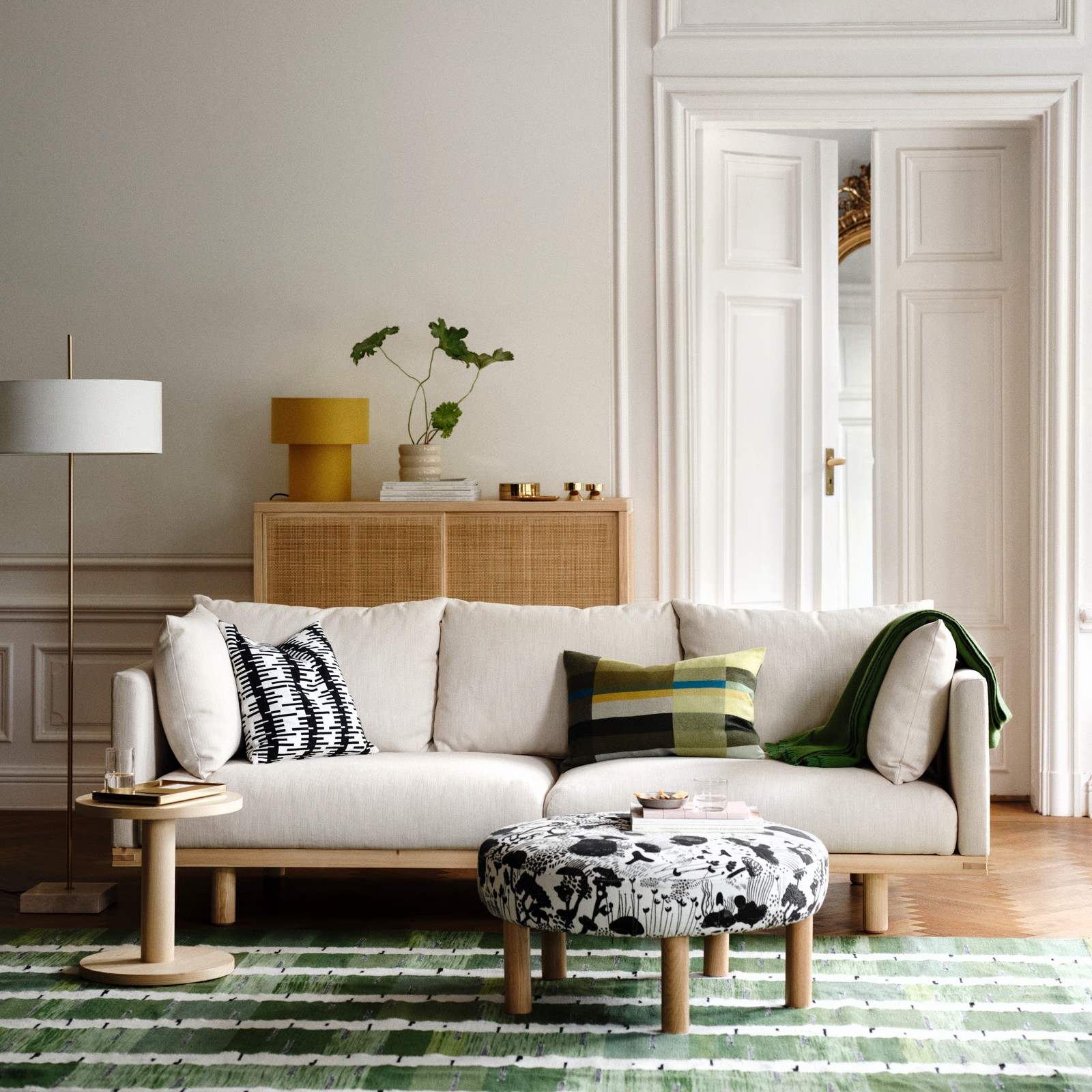 Image of a white living room with molding on the walls. There is an off-white and wooden sofa with a black and white patterned ottoman in front. On the sofa are patterned pillows, and there is a rattan console table on the wall behind the sofa with a yellow upholstered lamp.