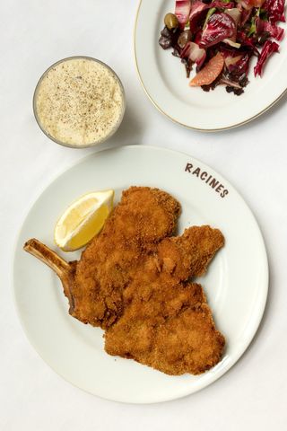 A veal cotoletta is served in a white, customized plate reading "Racines" in dark red, along with a radicchio salad and a dip, all placed on a white tablecloth.