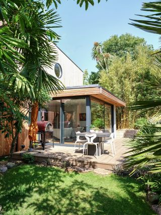 A modern home glass extension with decking area surrounded by palm trees and green lawn