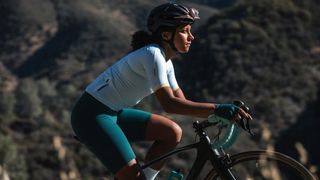 A woman of colour with afro hair cycles her road bike from left to right of the image, wearing green and blue Velocio road kit