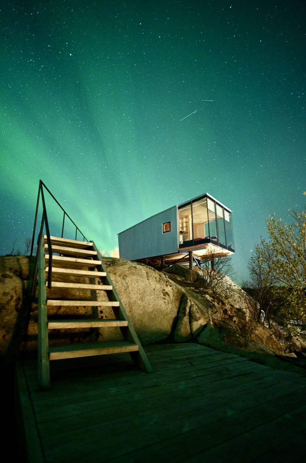 A room at hotel Manshausen in Lofoten Islands with the Northern Lights in the sky at night. Streaks of green light spread across the sky with stars. A ladder is pictured in the foreground leading up to the modular hotel room that is cantilevered over a small cliff.