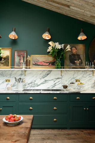 A bottle green kitchen with marble backsplash and countertop, brass handles and vintage framed oil paintings lining a wall shelf.