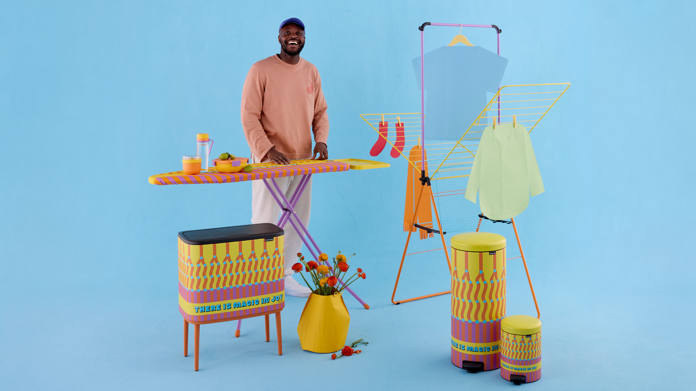 Yinka Ilori x Brabantia collection against blue background. Yinka is stood smiling behind the ironing board.