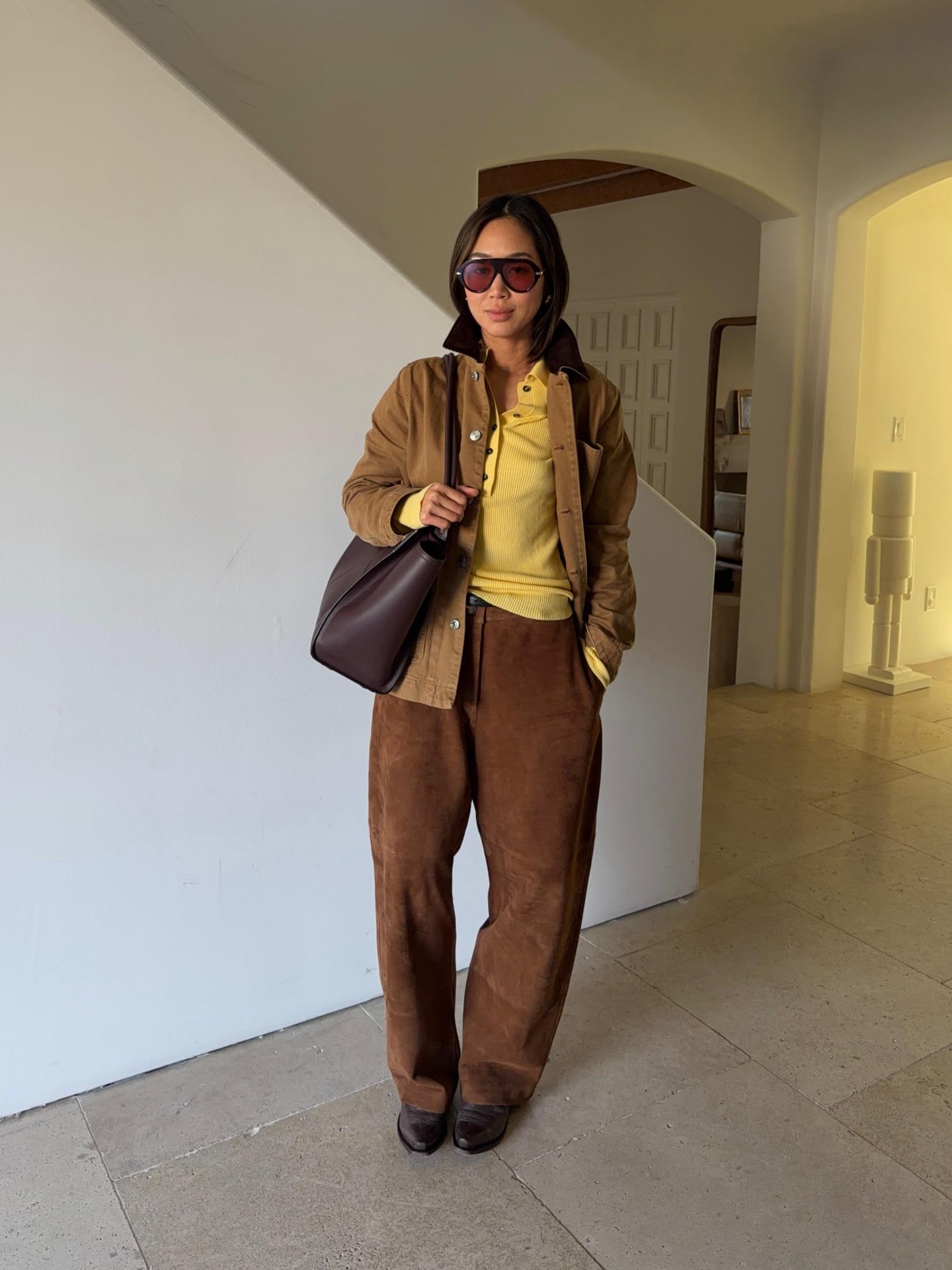 Fashion person Aimee Song wears a transitional winter to spring outfits 2026. She wears a bright yellow polo, suede brown trousers and brown loafers and a brown barn jacket. She has a dark brown tote over her shoulders and has oversized orange lens sunglasses on. She is stood in front of a white staircase looing at the camera in a recent image from her Instagram.