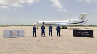 four people in blue flight suits stand on a tarmac in front of a small jet aircraft