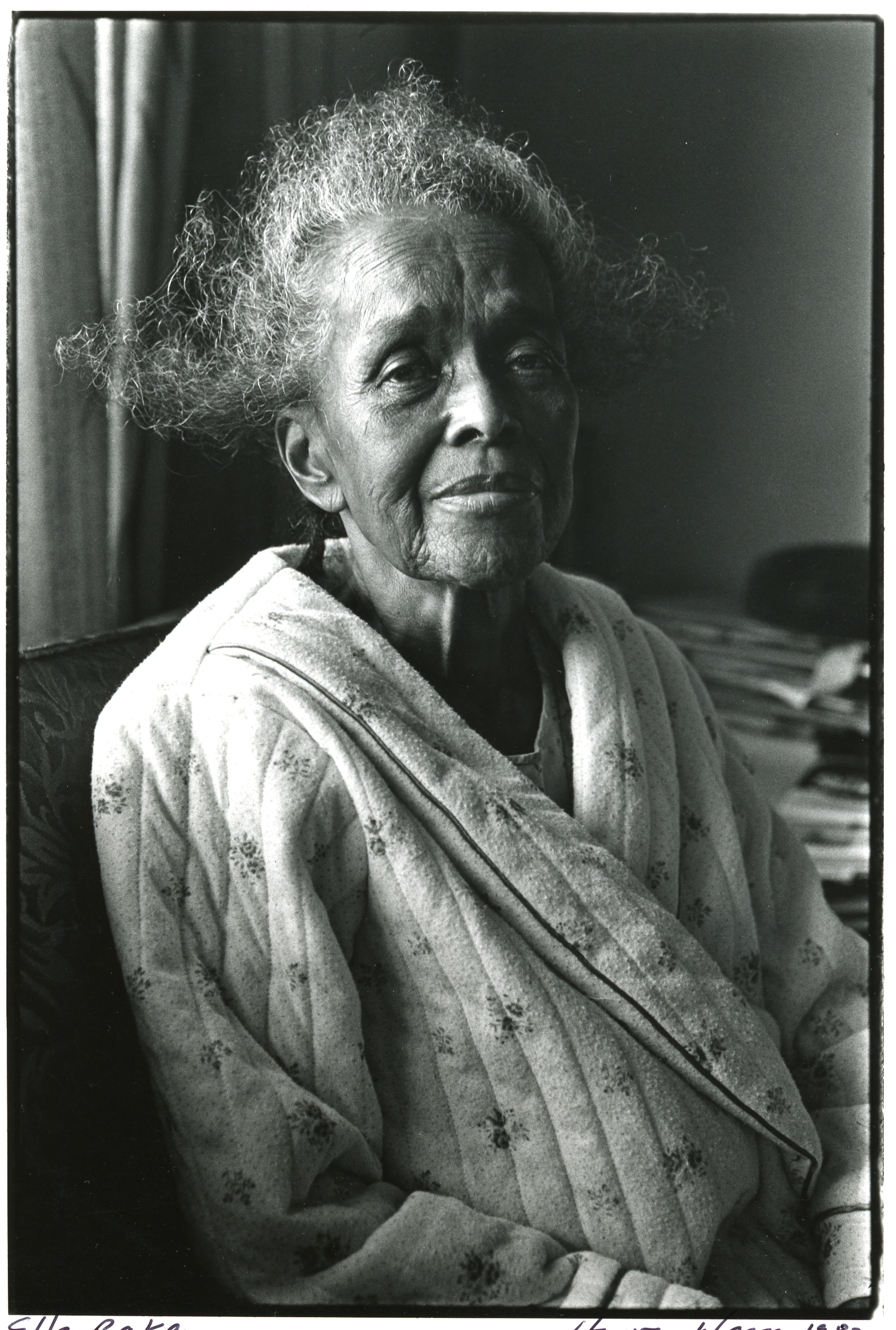A portrait of an elderly Ella Baker with curly grey hair, wearing a quilted floral robe and smiling softly at the camera