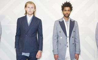 Two guys wearing the Hardy Amies S/S 2015 collection. On the left the guy is wearing a white shirt and navy suit with a blue scarf. On the right the guy is wearing a gray shirt, white jacket, white pants and a navy overcoat