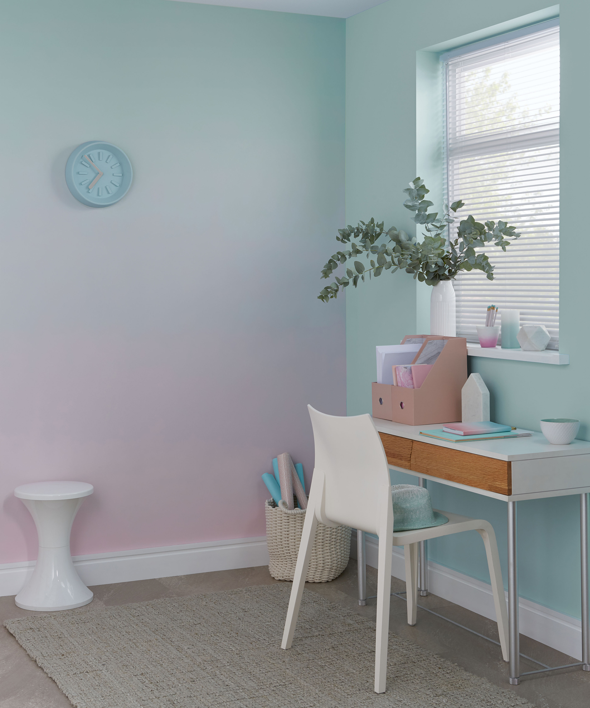 Pink and green ombre wall decor in home office, with small white desk and chair, and a storage basket in the corner and small white side table