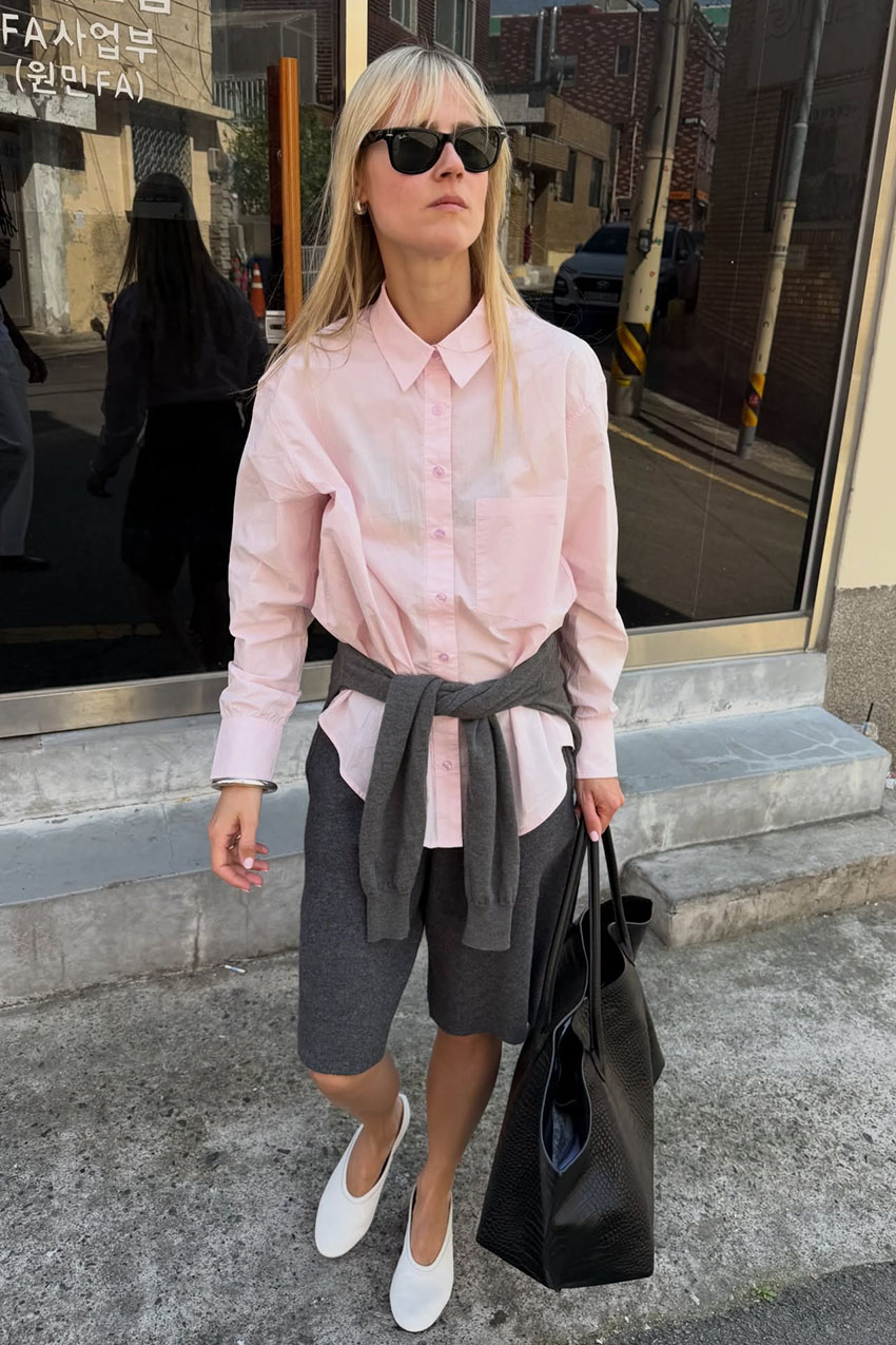 a woman wearing a baby pink button-down, gray shorts, white flats, and black leather bag