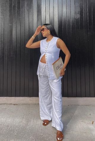 A woman wearing brown sunglasses, gold earrings, a white vest, a Celine clutch, white linen pants, and tan slides.