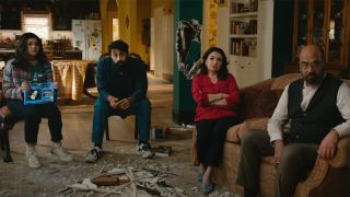 Kamala Khan, aka Ms. Marvel, sitting with her family in their living room. There is a pile of rumble on the floor between them all. From left to right: Kamala Khan (teen girl, long dark hair, is holding a holographic tablet in her lap), male relative (slightly older, short dark hair and a big moustache and beard), Kamala's mother (long dark hair, sitting with arms crossed), and Kamela's father (balding man, wearing glasses and has a beard and moustache).