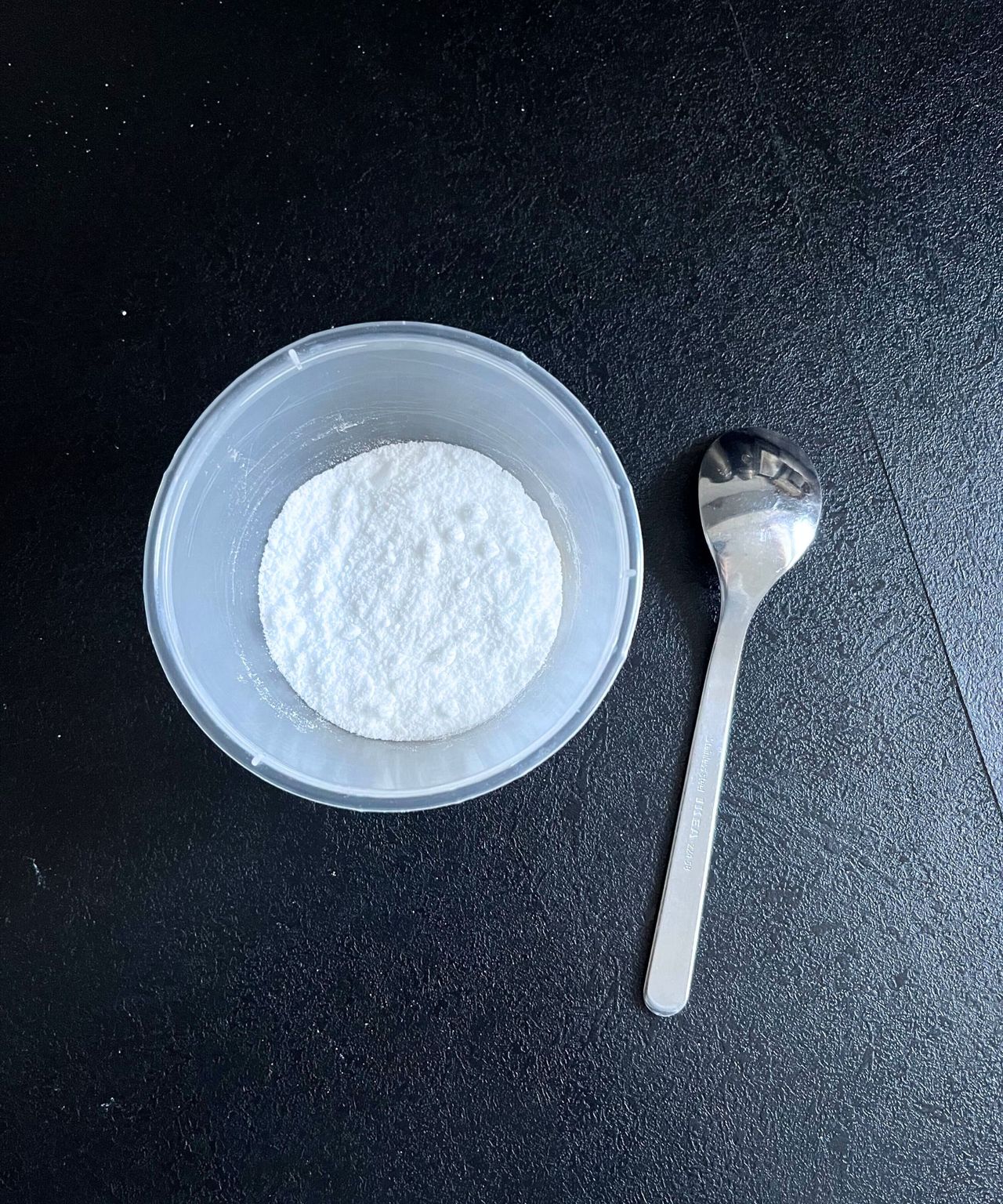 A round plastic container with baking soda inside. A silver teaspoon beside it.