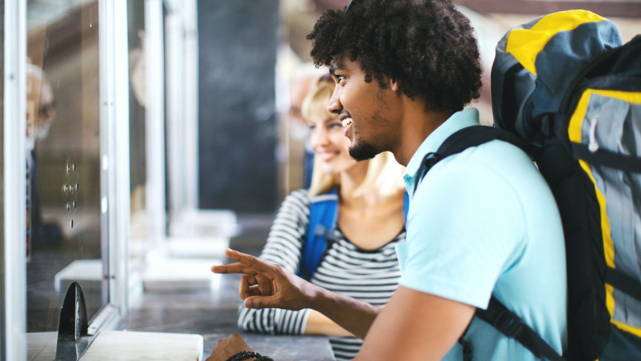 Young couple wearing large backpacks buying travel tickets at ticket counter