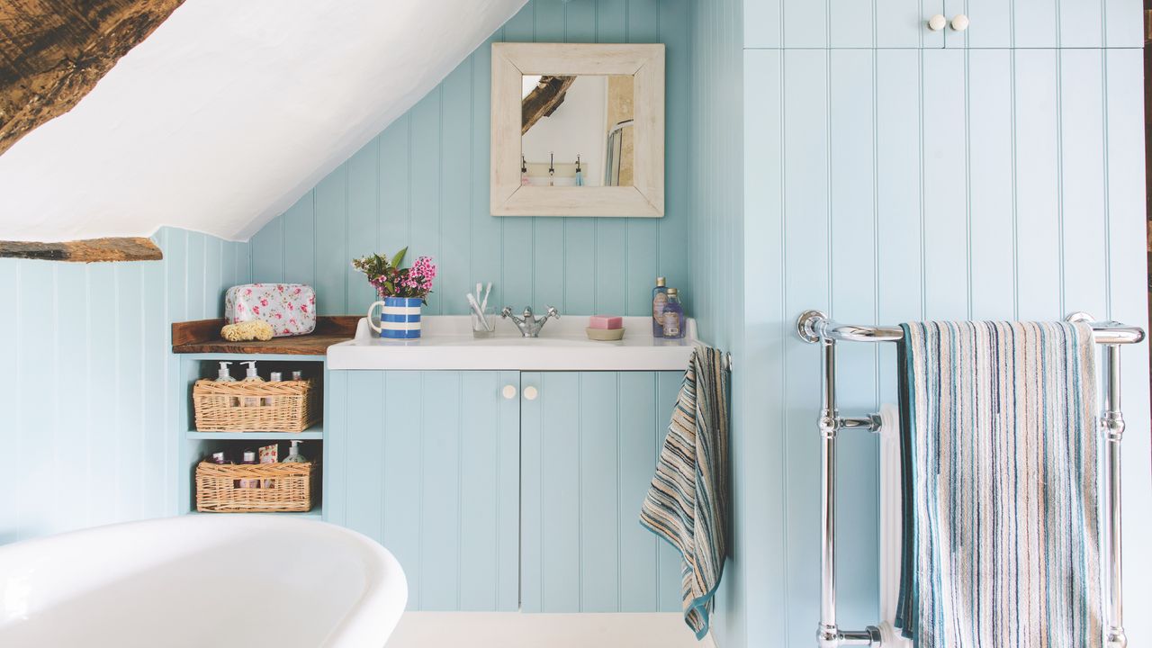 coastal bathroom in loft with pale blue tongue and groove, white ceiling, bath and floor, mirror, storage baskets, radiator, striped towels, beams 