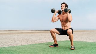 Man performing dumbbell thrusters outdoors on grass with dumbbells on shoulders