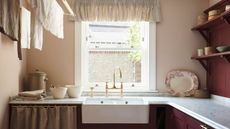 Neutral laundry room with bright window, butler sink, ceiling mounted drying rack, and dark red wooden cabinets and wall panels. Marble worktops and brass hardware finish the cottage look