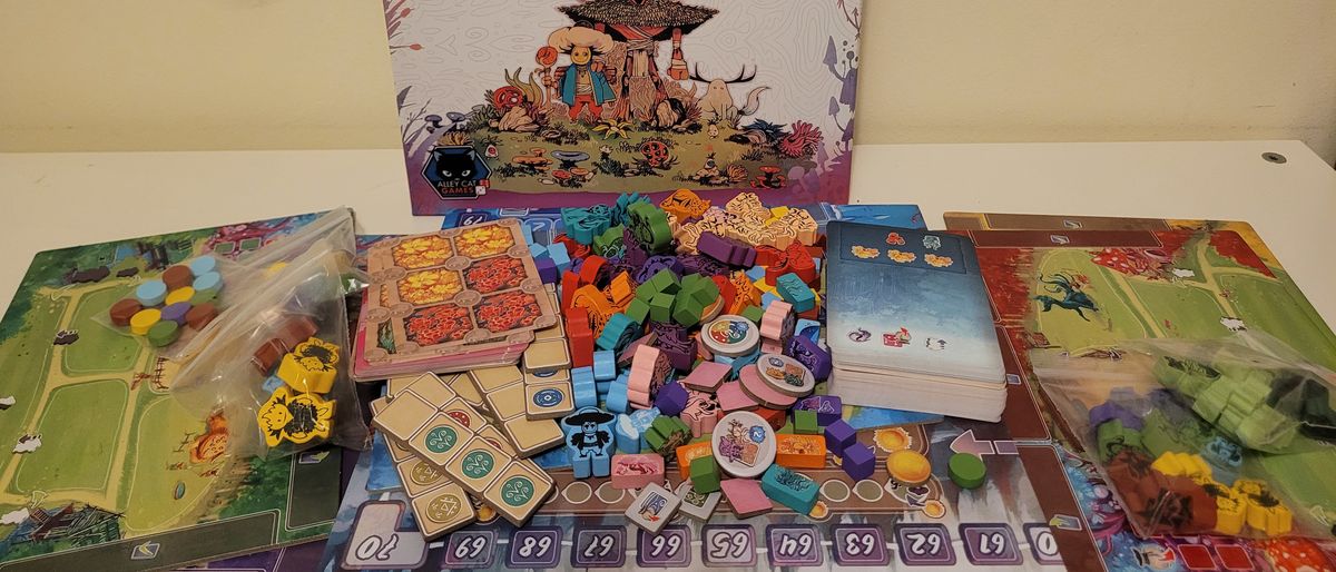 Arborea box, tokens, meeples, and boards on a white surface, against a cream-colored wall