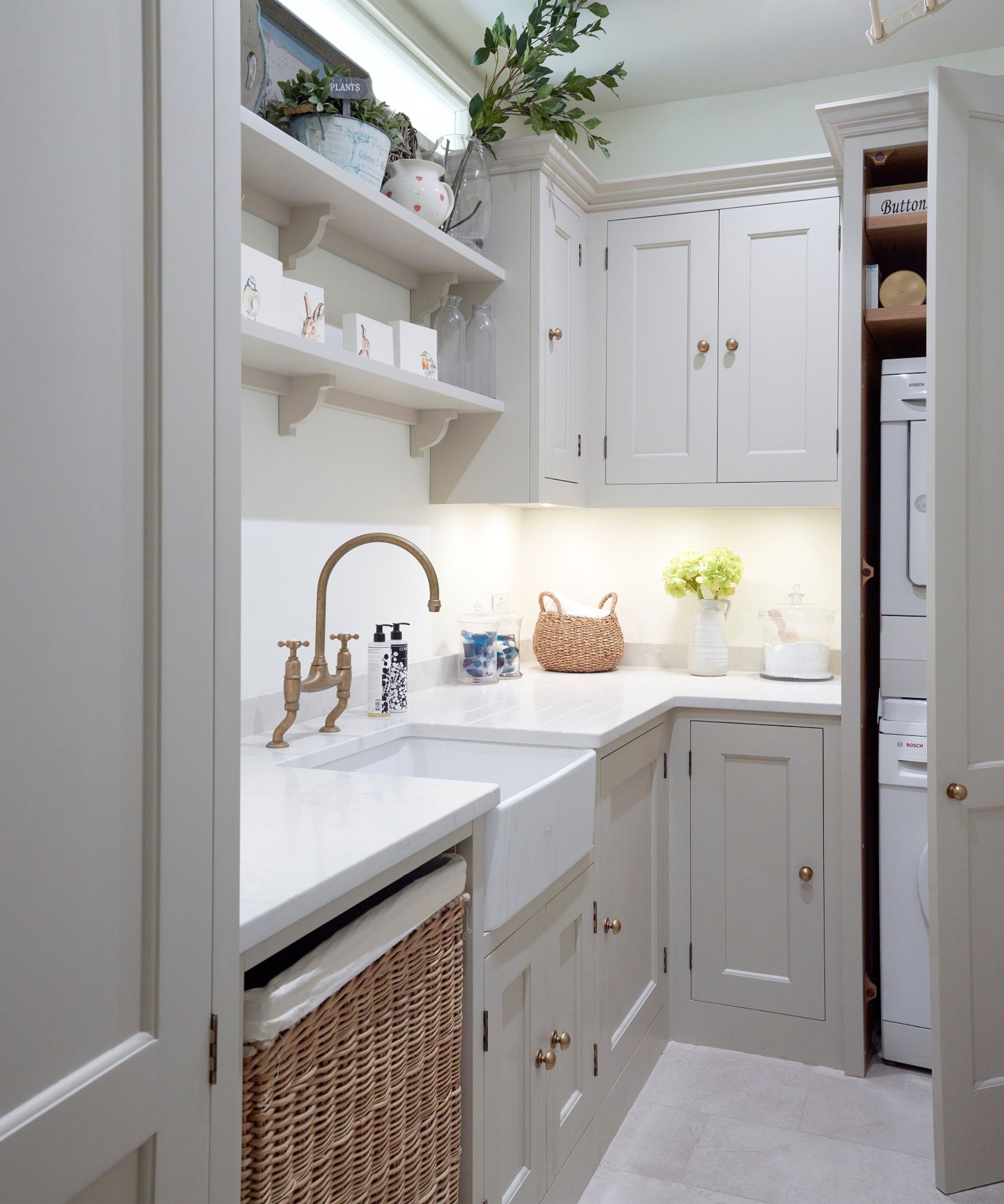 A compact grey utility room with a Belfast sink, brass bridge tap, white marble worktop, open shelving with plants and ceramics, wicker laundry basket below.