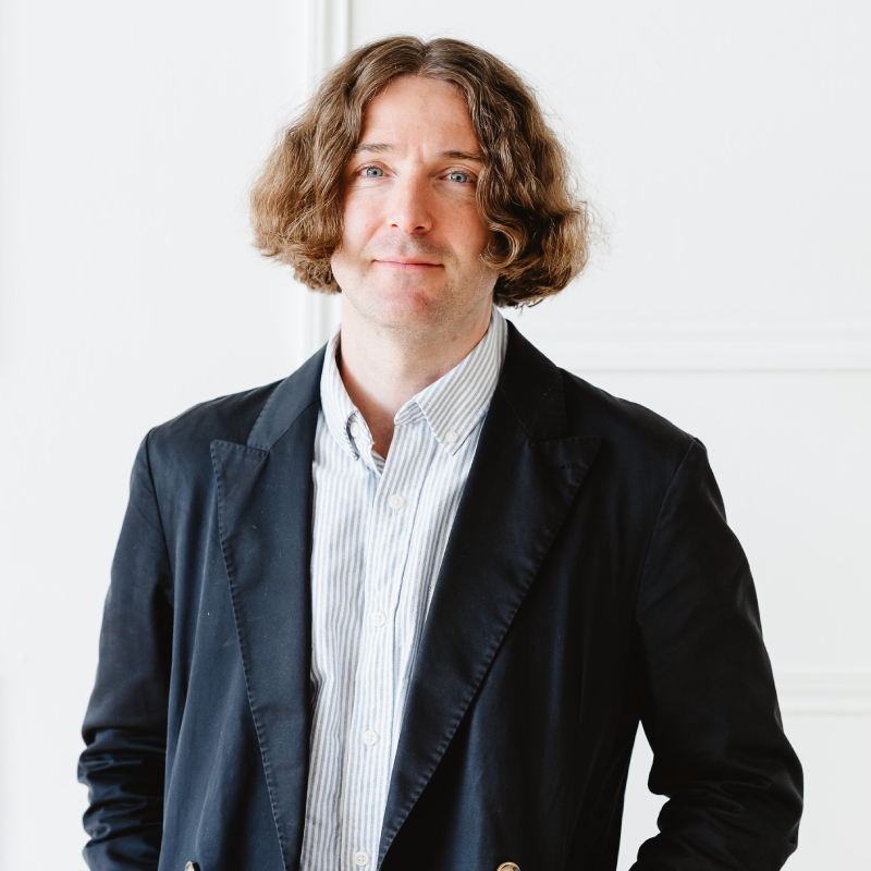 male with long brown hair wearing blue blazer and striped shirt