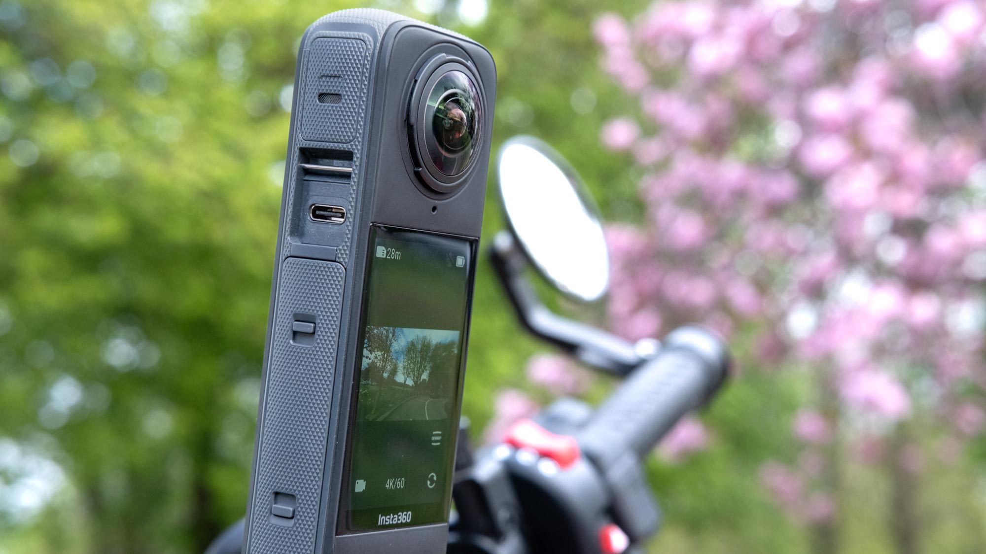 A photo of the Insta360 X4 mounted on motorcycle handlebars with green and pink foliage in the background. On the screen of the camera is the same background scene. The USB-C cover has been taken off to show the connection port.