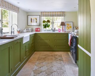Kitchen with green kitchen cupboards with two windows with patterned blinds, stone flooring with textured rug, countertops styled with ornaments and accessories