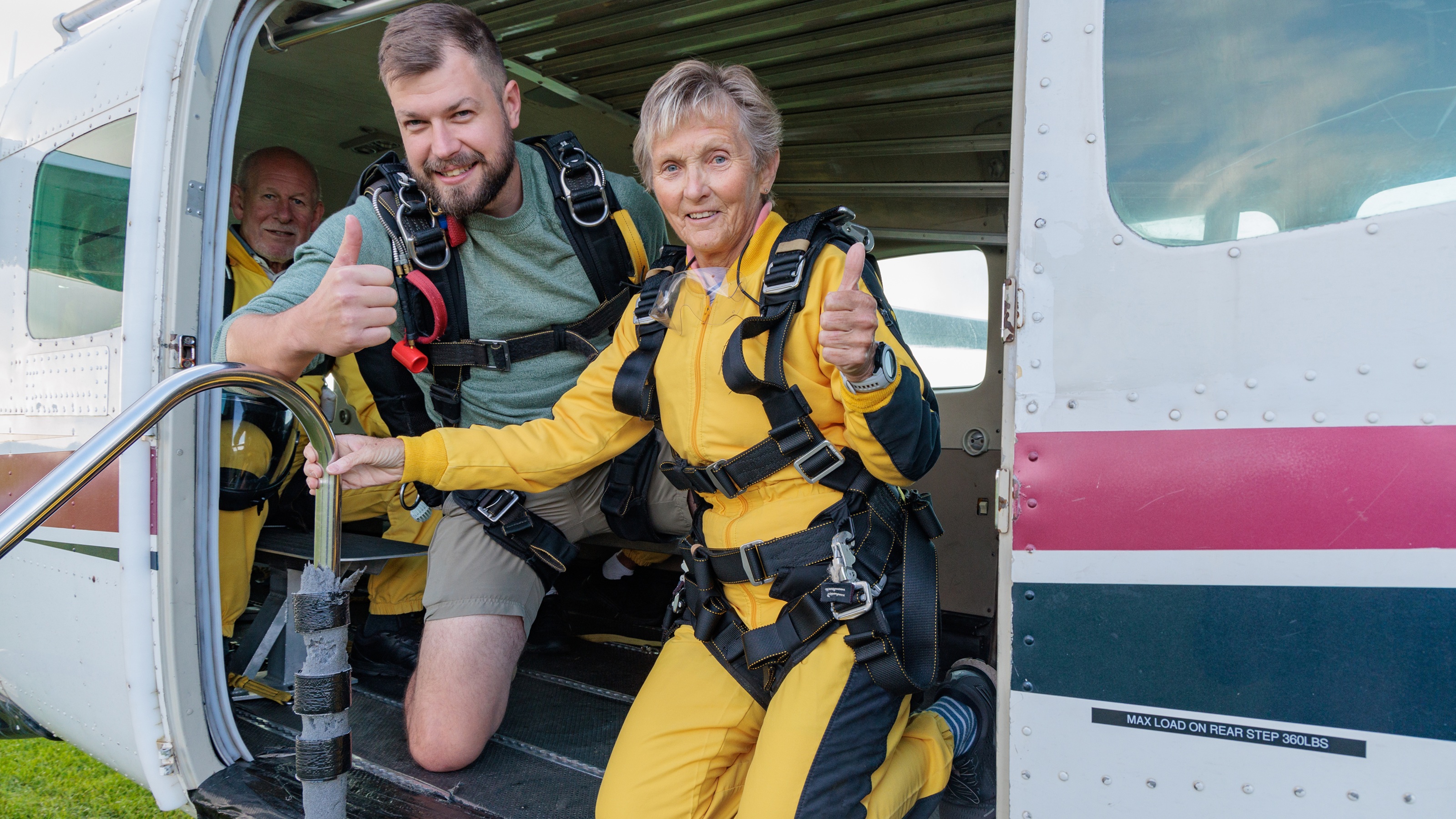 An older skydiver gives a thumbs-up before heading up on an airplane with her instructor.