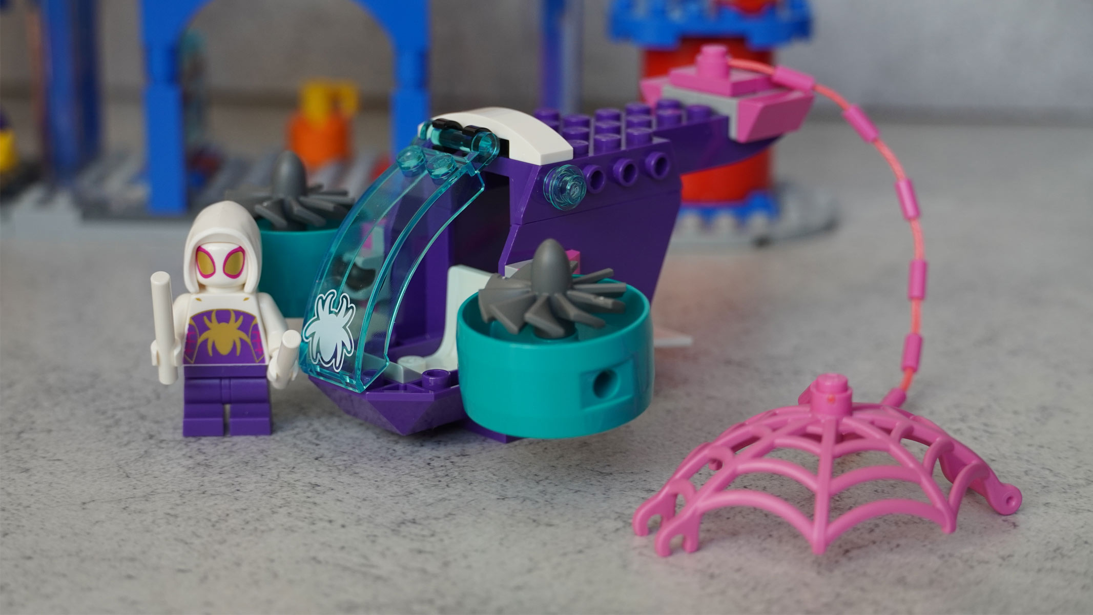 A pink web is attached to a purple helicopter with a minifigure of someone with a white hood and purple body standing next to it.