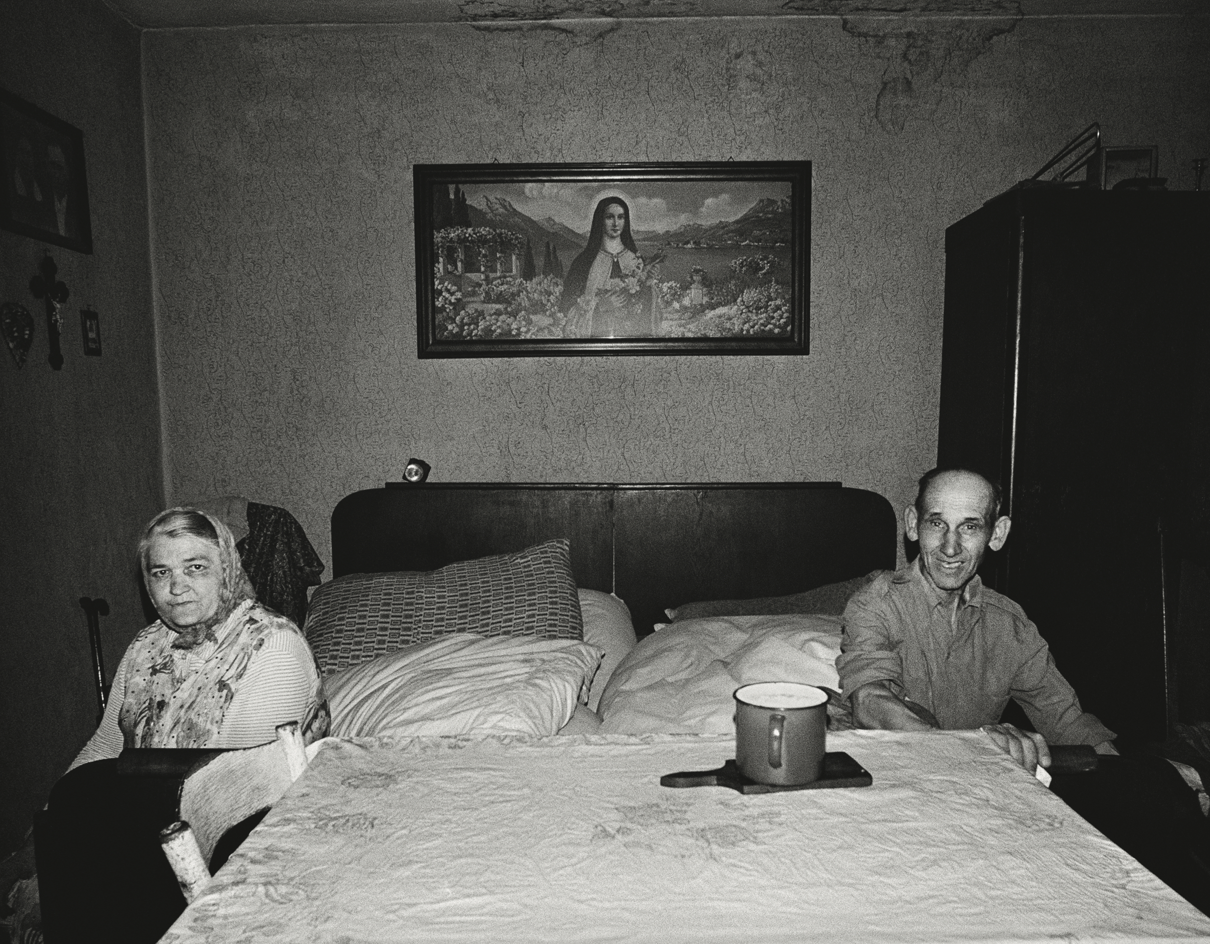 Black and white photograph of anelderly couple sat on either side of a bed. Hanging upon the wall between them is a religious picture of Jesus.