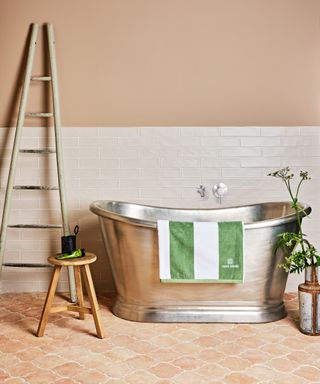 silver roll top bath on terracotta floor tiles with wooden ladder, white wall tiles and terracotta painted wall above