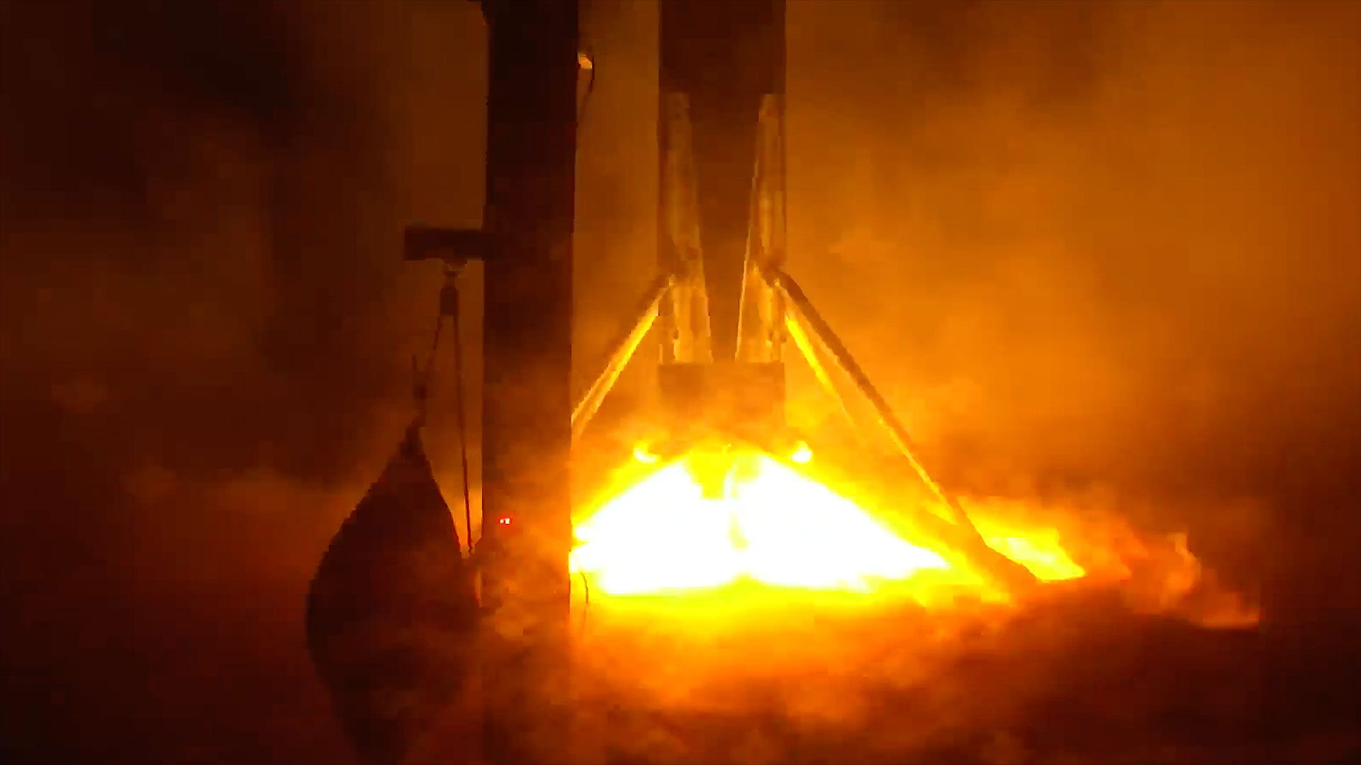 the bright orange and white flame from a rocket engine lights the scene at night of a booster stage landing on an ocean-based platform