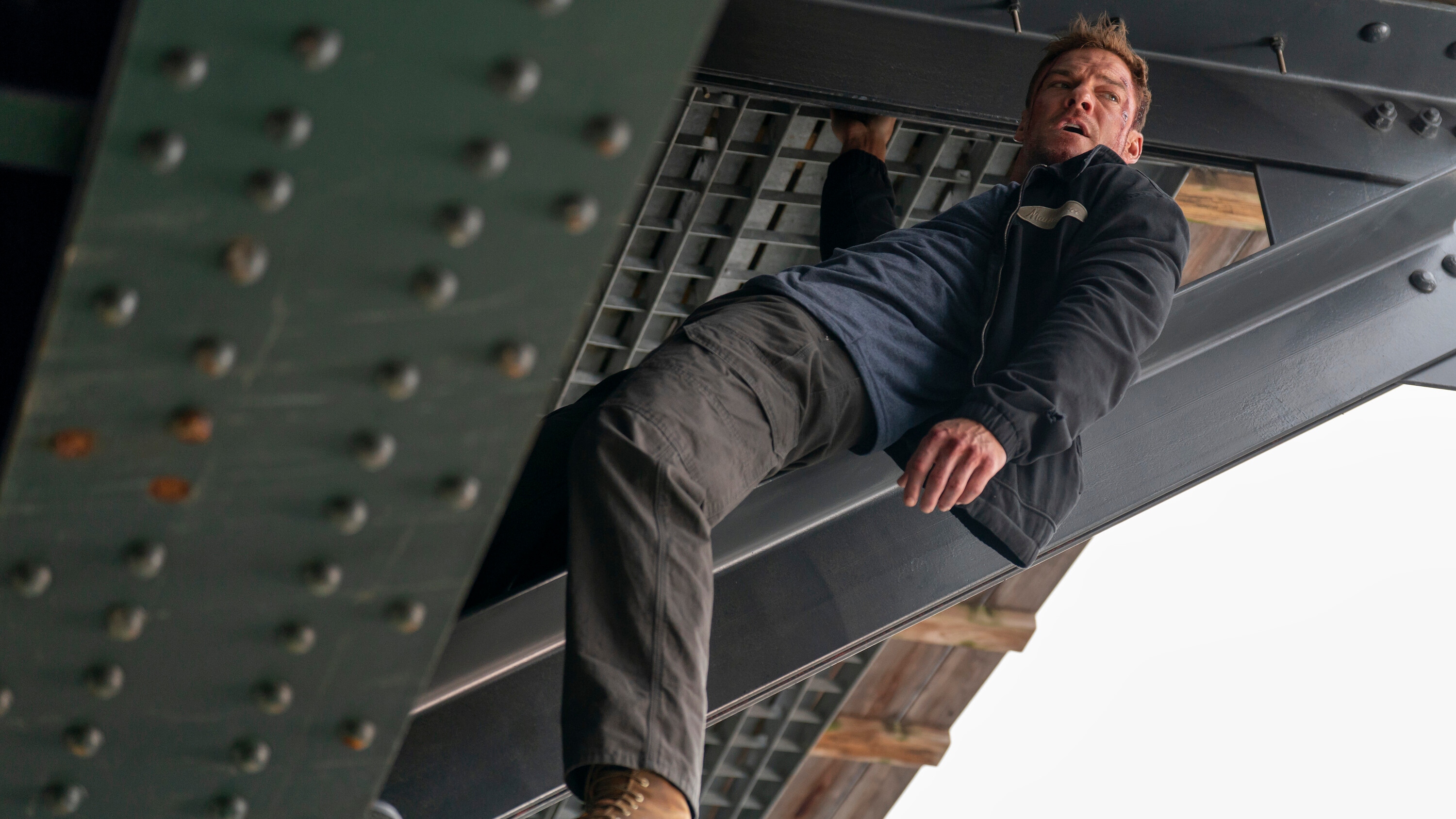 Alan Ritchson as Jack Reacher with facial injuries hanging off a bridge.